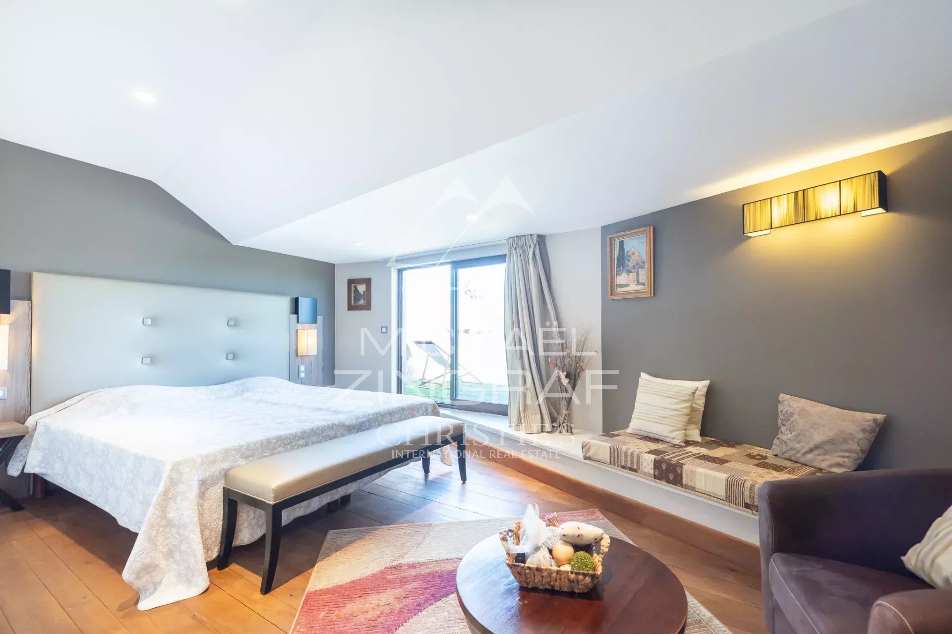 Spacious hotel bedroom with a king bed and padded headboard, a bench at the foot, window seating, and a round table with a basket of snacks nearby.
