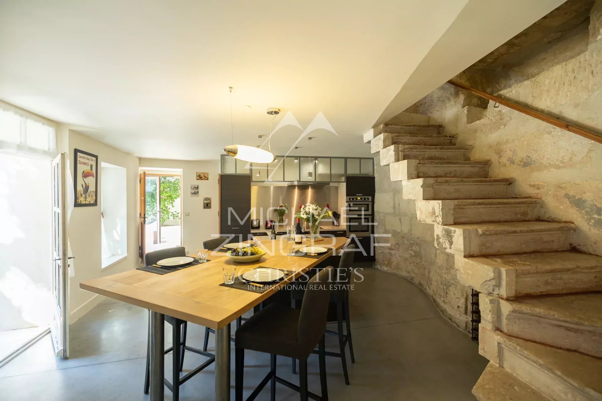 Open-plan kitchen and dining area with a wooden table, stone-wall staircase on the right, and stainless steel kitchen in the background.