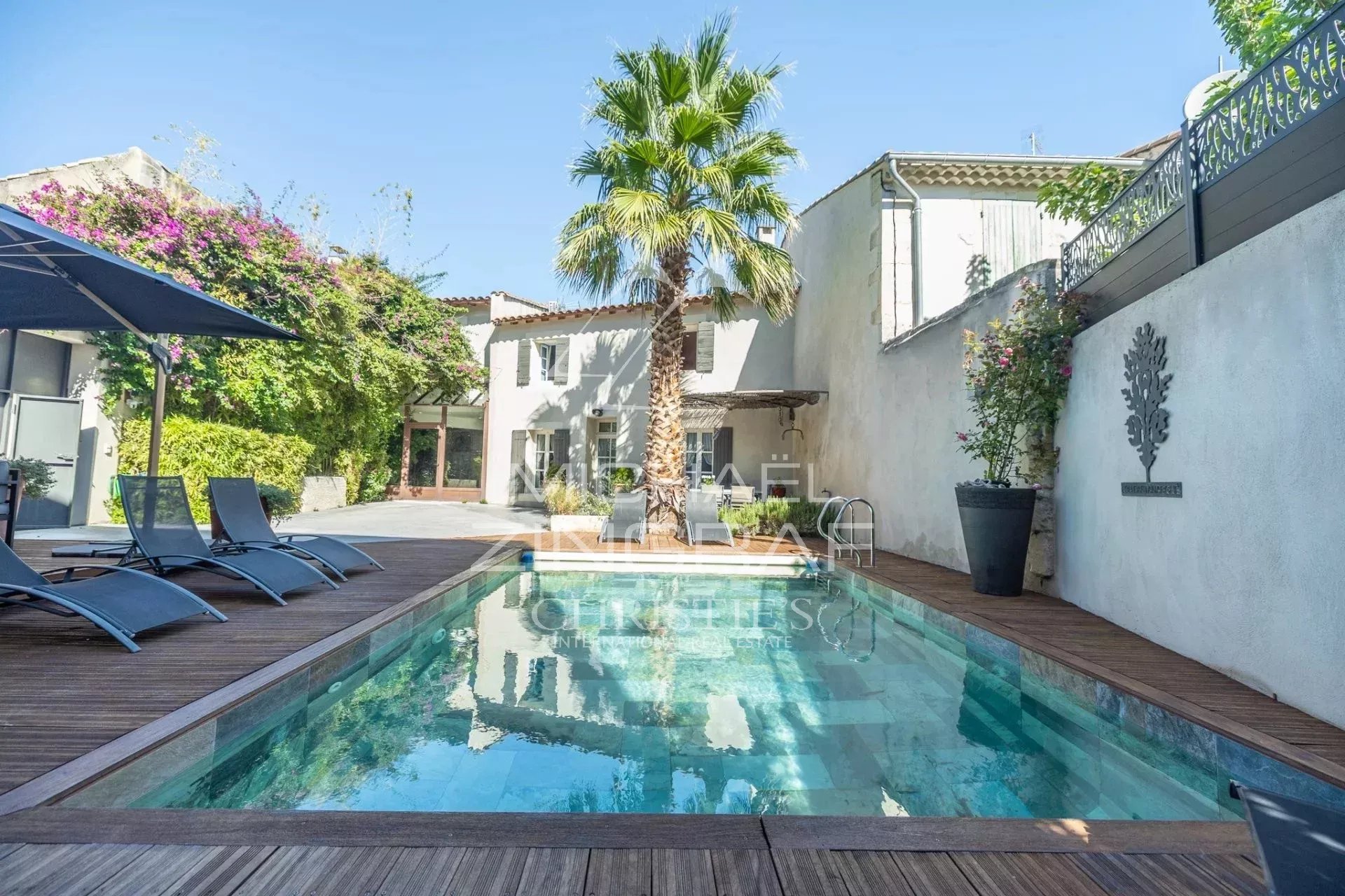 Poolside backyard with a rectangular pool, wooden deck, palm tree, and blue lounge chairs under a sunny sky.
