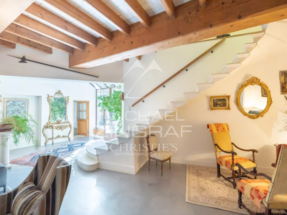 Spacious living area with white walls, exposed wooden beams, and a white staircase; vintage gold mirrors and ornate furniture, plants, and a striped chair nearby.