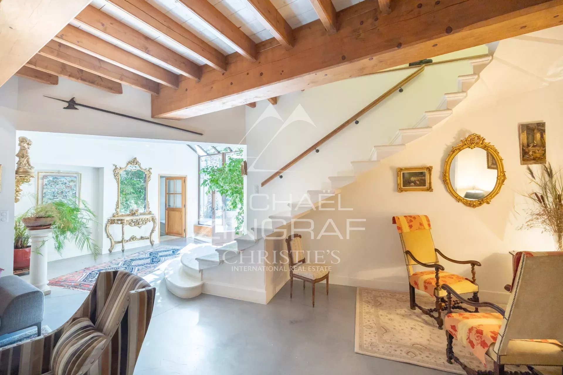 Spacious living area with white walls, exposed wooden beams, and a white staircase; vintage gold mirrors and ornate furniture, plants, and a striped chair nearby.