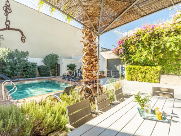 Outdoor backyard with a rectangular pool, wooden deck, and a shaded dining table under a thatched canopy; plants and lounge chairs along the wall behind.