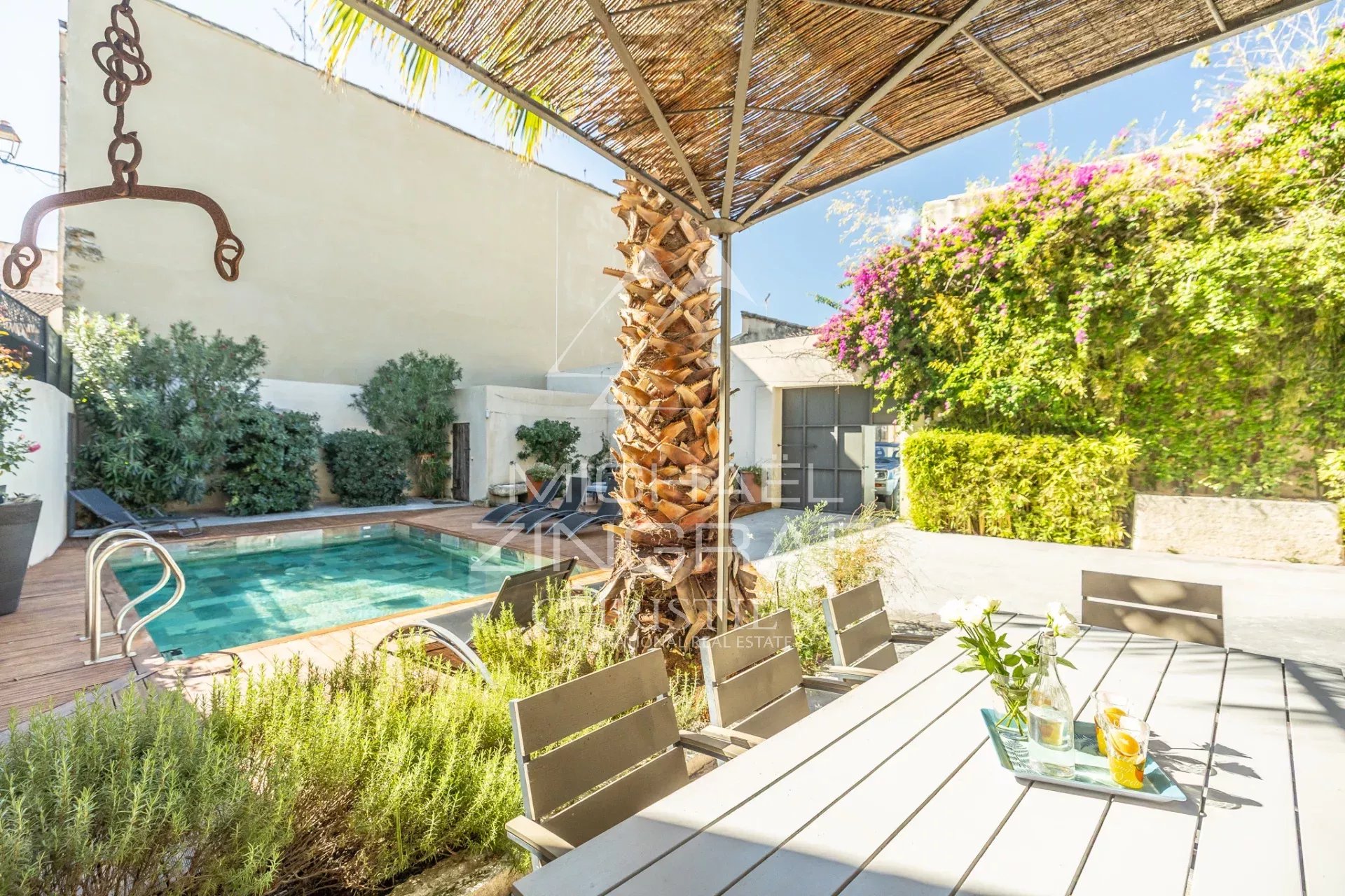 Outdoor backyard with a rectangular pool, wooden deck, and a shaded dining table under a thatched canopy; plants and lounge chairs along the wall behind.