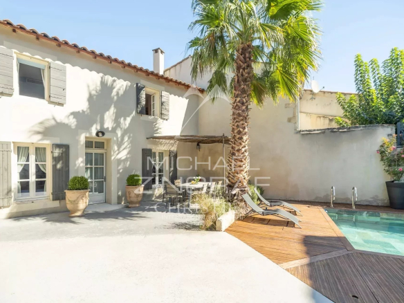 Sunny courtyard of a white stucco house with gray shutters, a tall palm tree, and a wooden pool deck with lounge chairs.