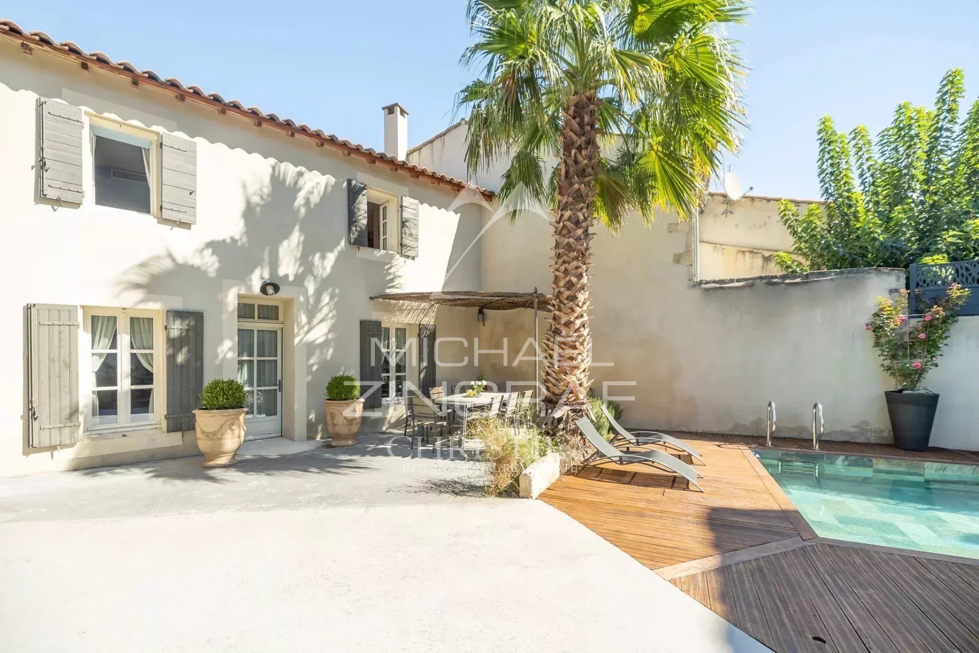 Sunny courtyard of a white stucco house with gray shutters, a tall palm tree, and a wooden pool deck with lounge chairs.