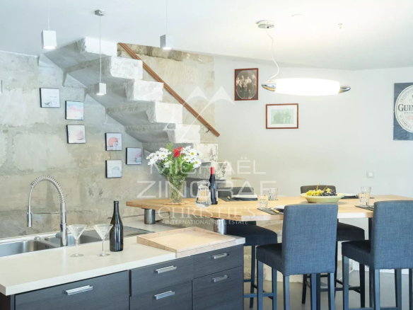 Modern dining area with a concrete wall, floating concrete staircase, and a wooden handrail; a wooden table set with a wine bottle, glassware, a fruit bowl, and a vase of flowers amid four blue-gray chairs.