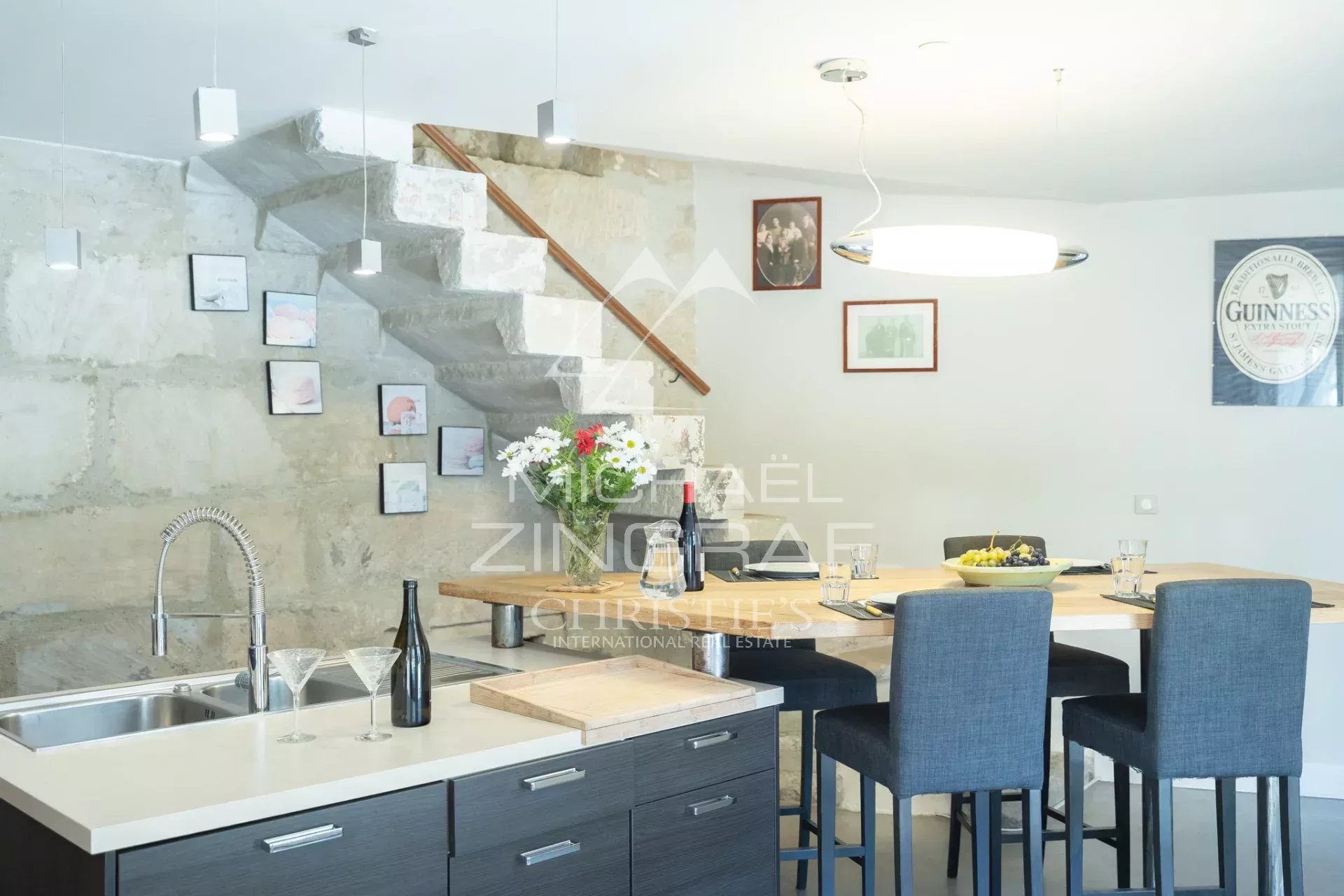 Modern dining area with a concrete wall, floating concrete staircase, and a wooden handrail; a wooden table set with a wine bottle, glassware, a fruit bowl, and a vase of flowers amid four blue-gray chairs.