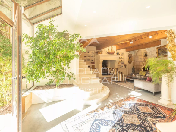 Bright living room with glass doors opening to a garden, large indoor plant, white fireplace, and a patterned rug under seating area with wooden beams above.