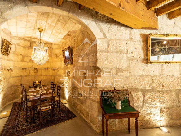 Stone-walled dining room with a vaulted arch, chandelier, and a long table set for six on a patterned rug, lit warmly by wall and floor lights.