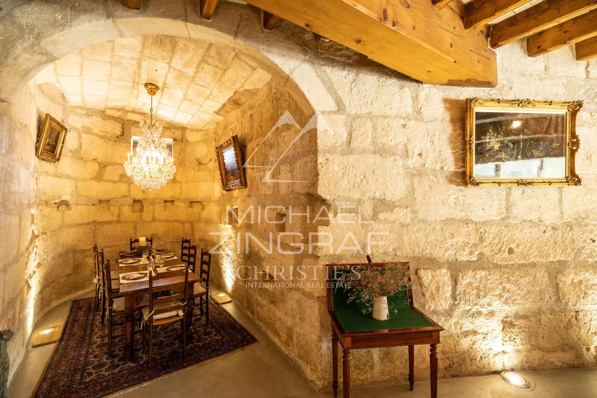Stone-walled dining room with a vaulted arch, chandelier, and a long table set for six on a patterned rug, lit warmly by wall and floor lights.