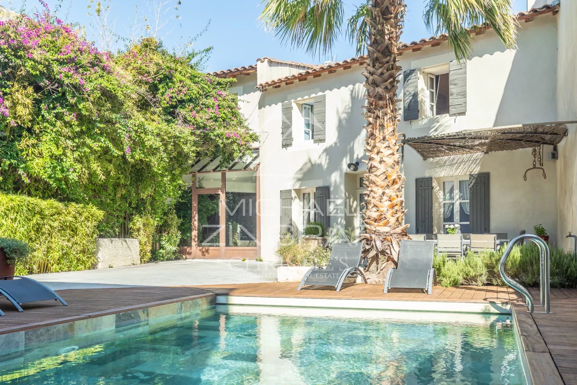 Villa with white walls, gray shutters, palm tree, and a swimming pool surrounded by a wooden deck and lounge chairs