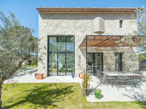 Stone-clad house with a glass-paneled door and large windows, outdoor dining table and chairs on a sunny patio, olive trees framing the scene