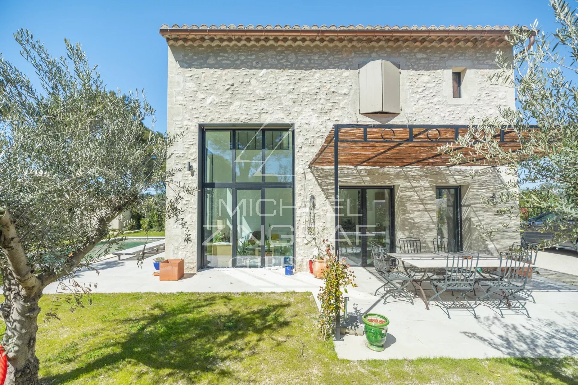 Stone-clad house with a glass-paneled door and large windows, outdoor dining table and chairs on a sunny patio, olive trees framing the scene