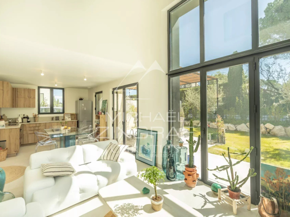 Bright open-plan living area with a white sofa, glass dining table, and a wooden kitchen ---- large windows reveal a sunny garden outside.