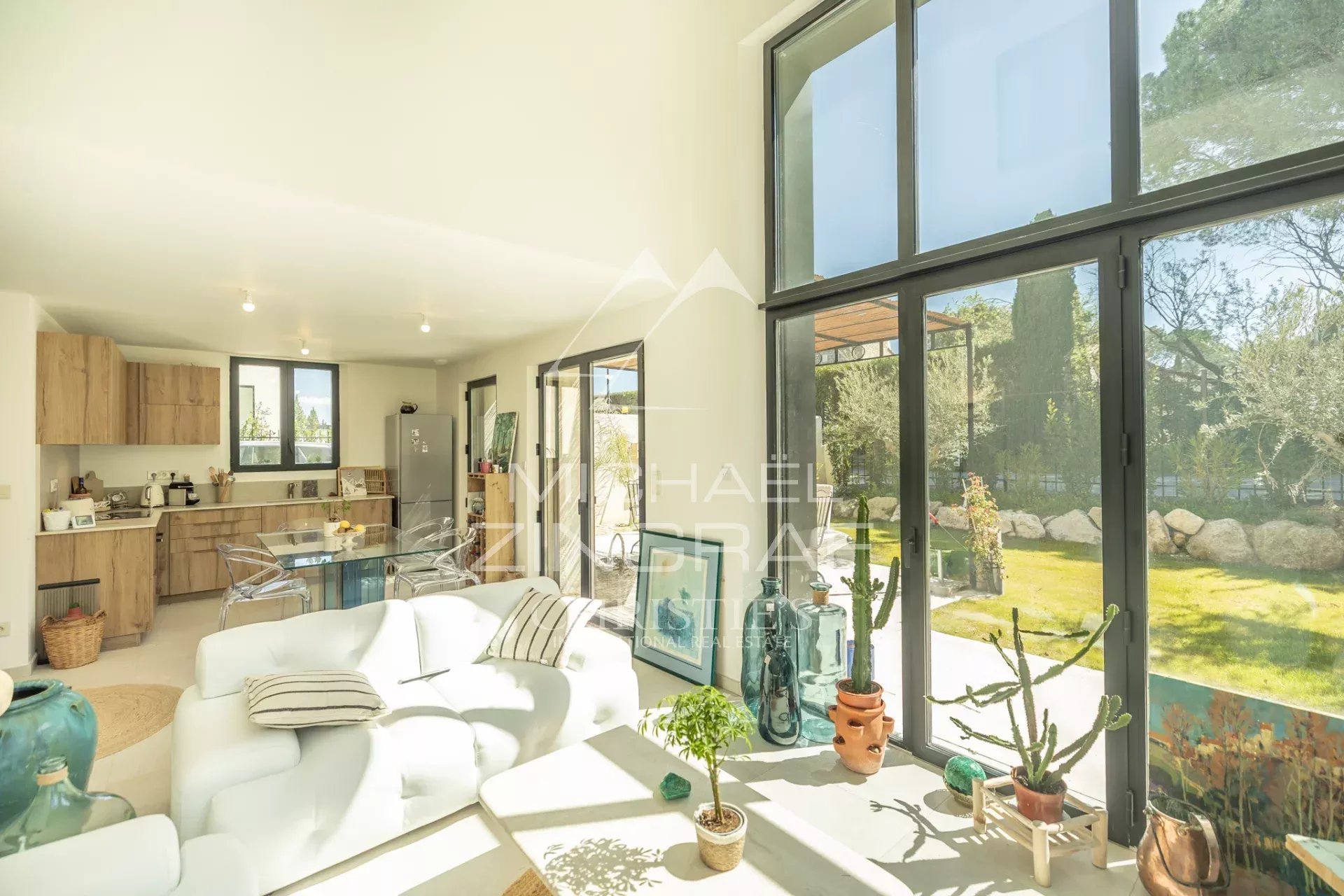 Bright open-plan living area with a white sofa, glass dining table, and a wooden kitchen ---- large windows reveal a sunny garden outside.
