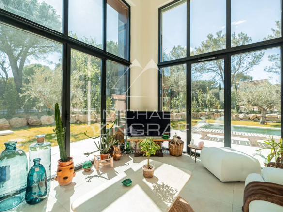 Bright modern living room with tall black-framed glass walls, several potted plants on a white coffee table, and a view of a yard with a pool outside.