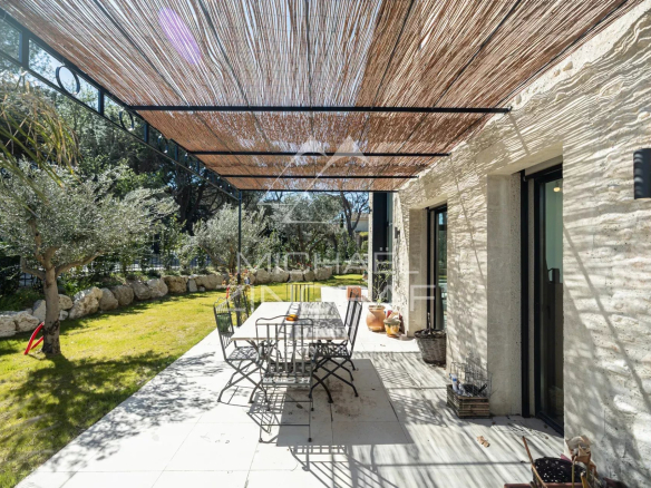 Outdoor patio with a long dining table and metal chairs under a woven reed shade, beside a stone house and olive trees.
