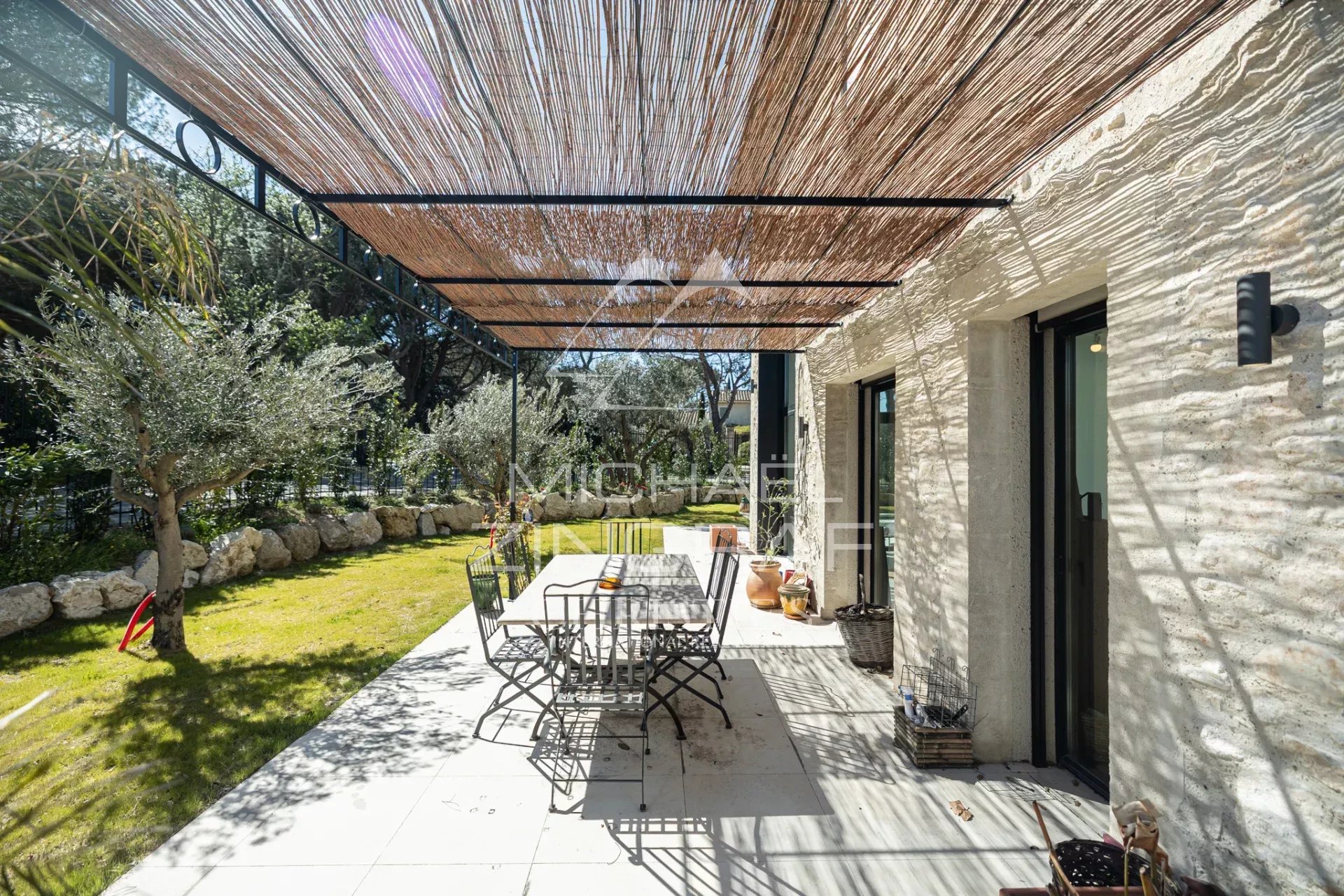 Outdoor patio with a long dining table and metal chairs under a woven reed shade, beside a stone house and olive trees.