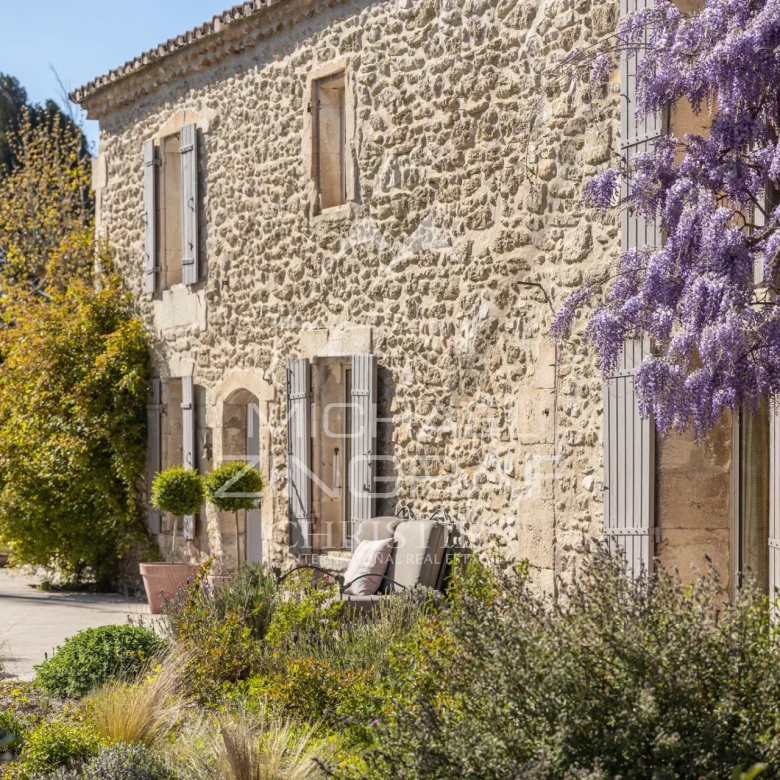 Stone house with shuttered windows, purple wisteria blooming along the wall, and a manicured garden.