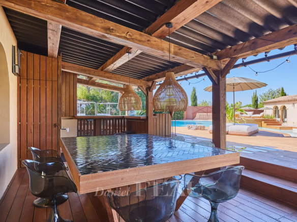 Terrasse en bois avec table et chaises, abritée sous une structure en bois, près d'une piscine.