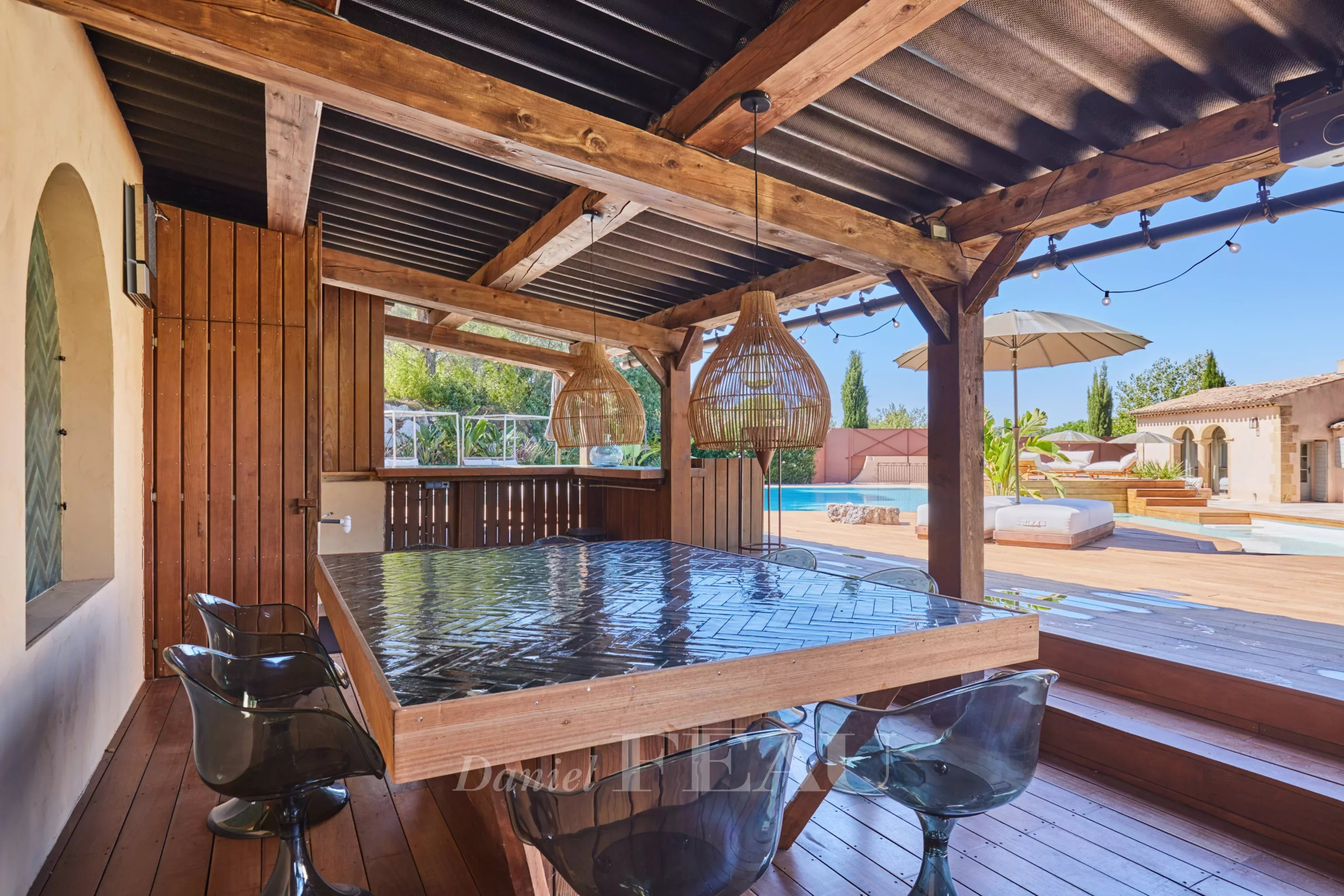 Terrasse en bois avec table et chaises, abritée sous une structure en bois, près d'une piscine.