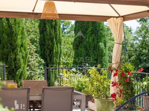 Shaded terrace with garden furniture and flowering plants in a wooded park.