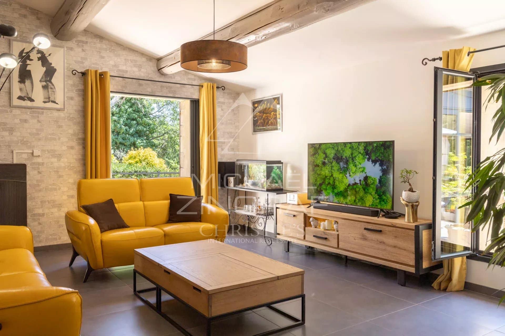 Interior of a bright living room with yellow sofas and a television, windows open onto the garden.