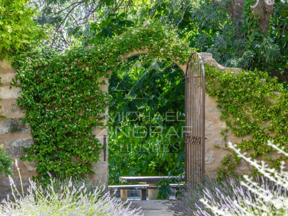 Entrance to a garden with a wrought iron gate, surrounded by greenery and flowers.