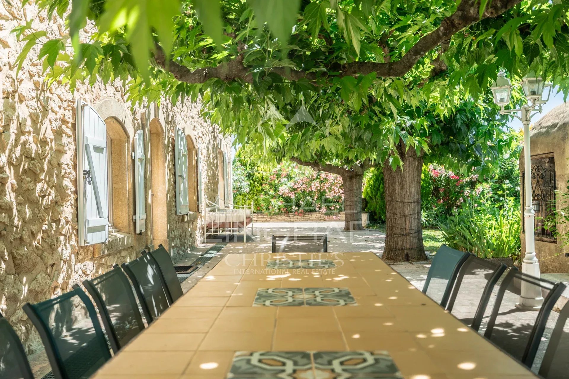 Terrasse d'un mas en pierre avec table et chaises sous un feuillage verdoyant.