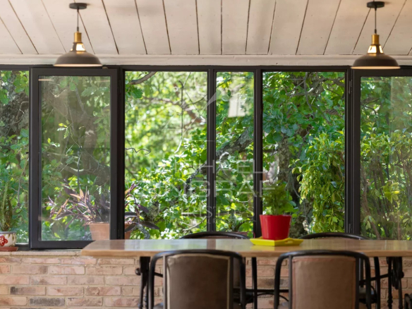Véranda lumineuse avec vue sur un parc arboré, mobilier en bois et plantes.