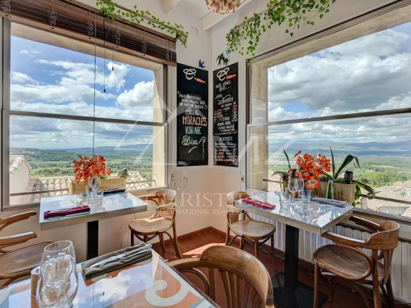 Intérieur d'un restaurant avec vue panoramique sur le Luberon, tables décorées et plantes.