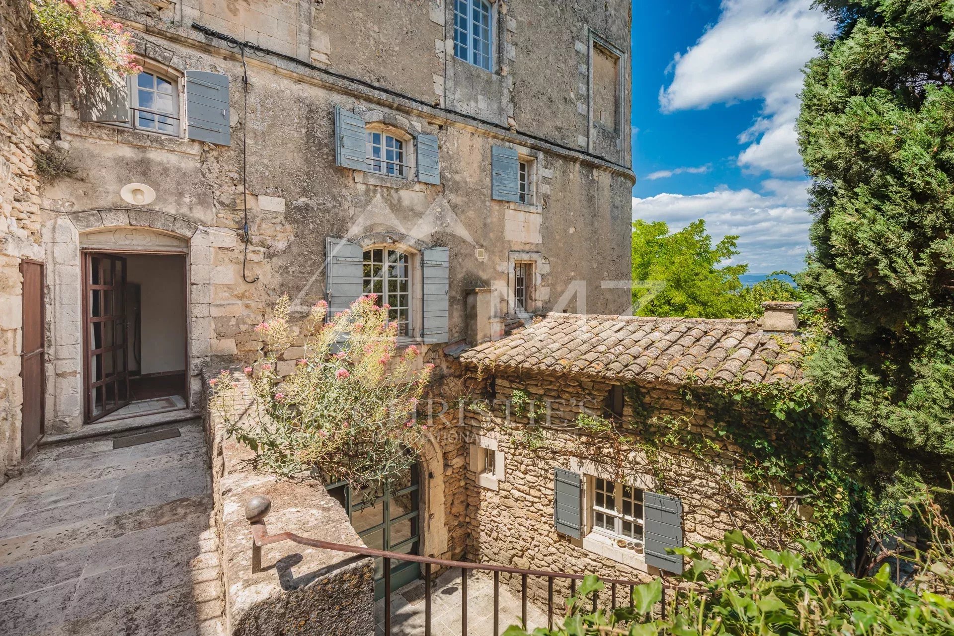 Entrance to a stone building with shuttered windows, surrounded by greenery in Ménerbes.