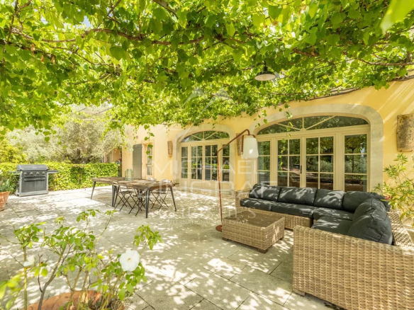 Terrasse ombragée d'une propriété provençale avec mobilier en osier et barbecue.