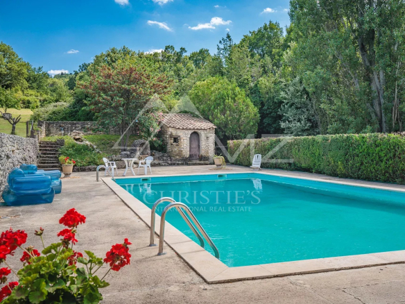 Piscine entourée de verdure dans un parc paysagé avec des arbres et un petit bâtiment en pierre.