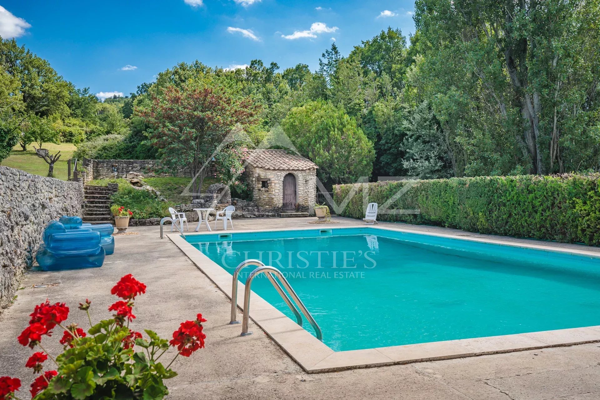 Piscine dans un parc paysagé Piscine entourée de verdure dans un parc paysagé avec des arbres et un petit bâtiment en pierre.