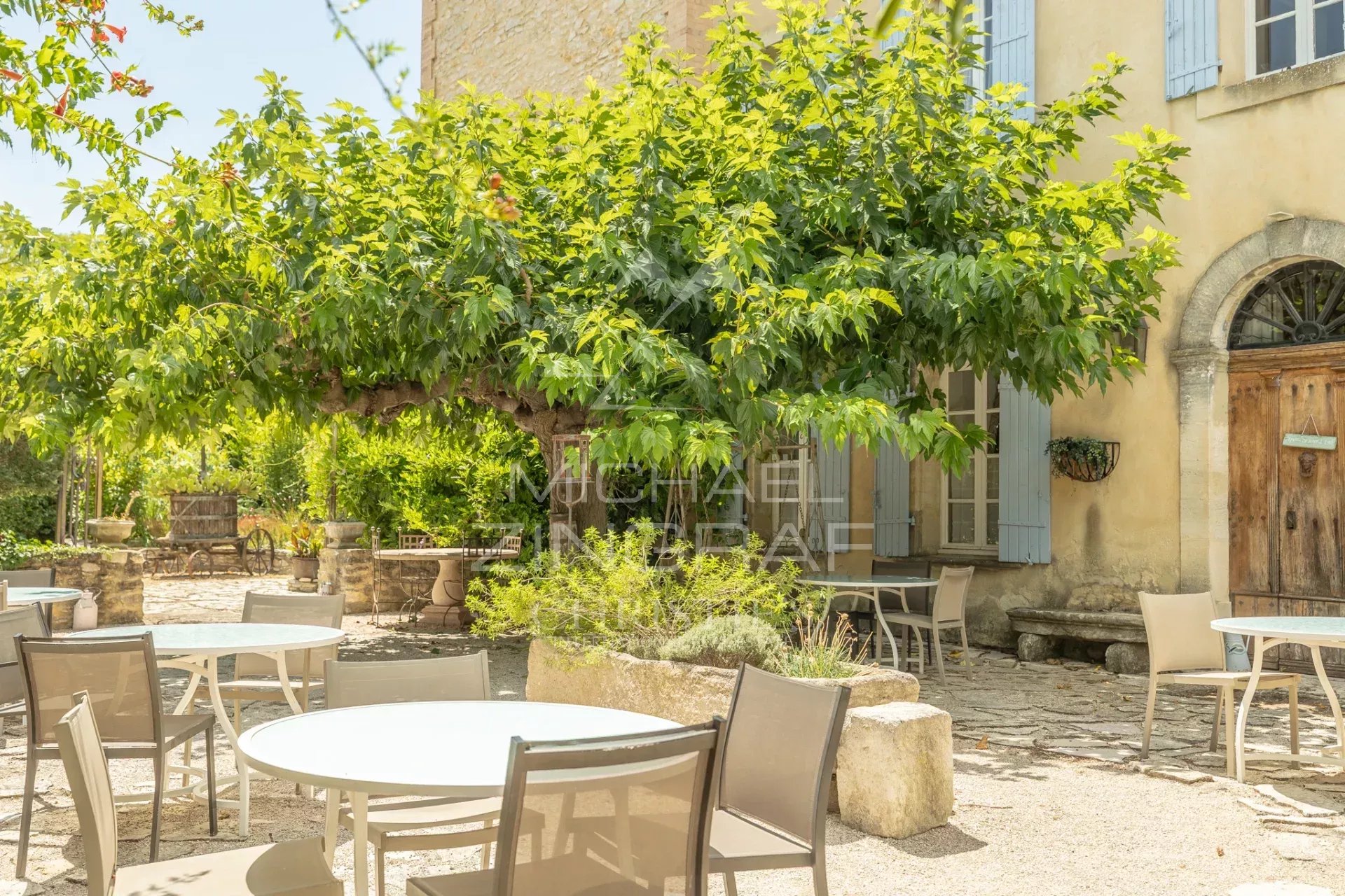 Terrasse d’un domaine provençal Terrasse avec tables et chaises sous un grand arbre dans un domaine provençal.