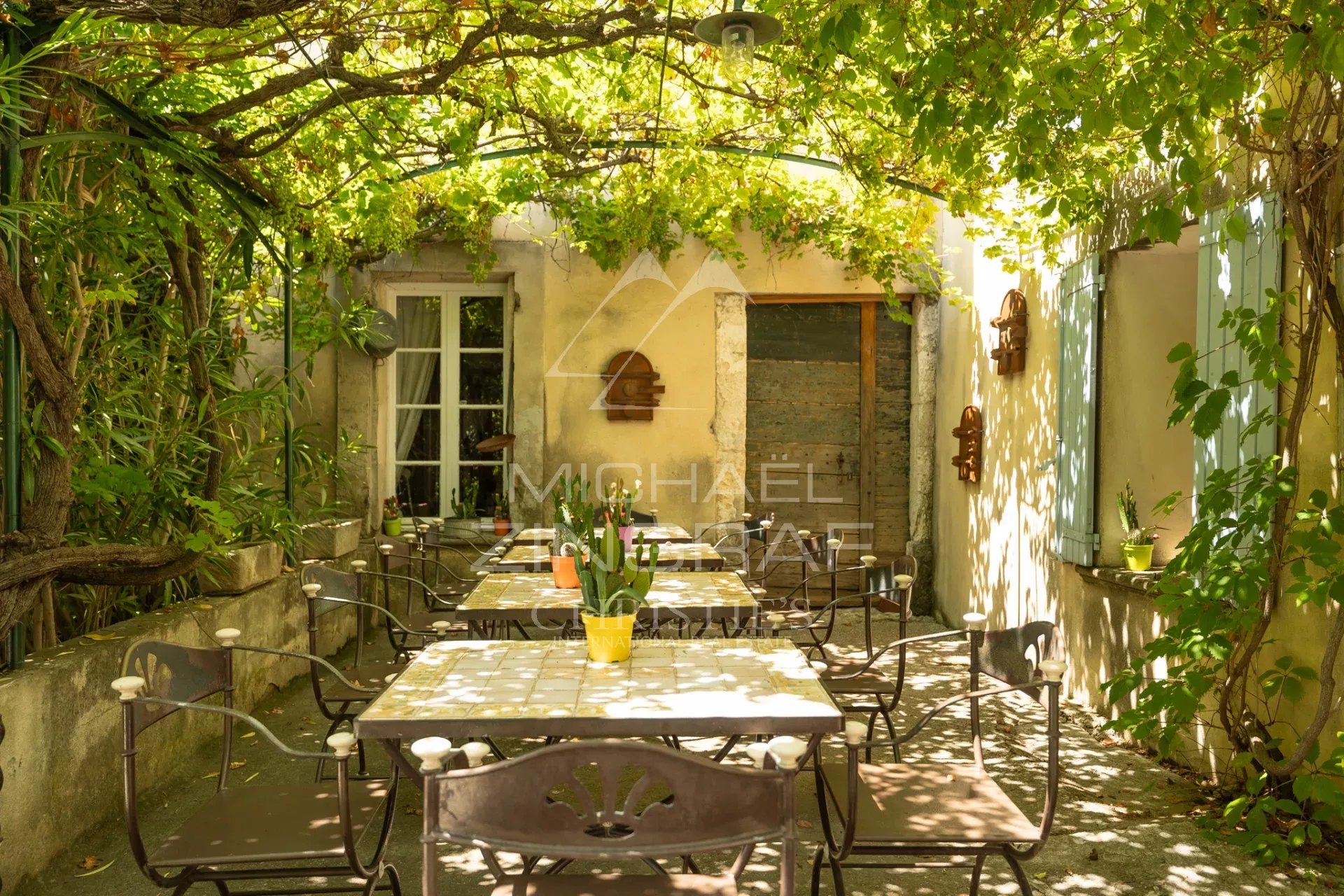 Terrasse d’un domaine provençal Terrasse ombragée avec tables et chaises, entourée de verdure dans un ancien prieuré.
