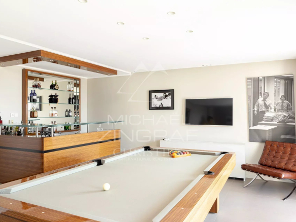 Contemporary home game room with a wooden bar, glass shelving of liquor bottles, and a pool table in the foreground. A brown leather lounge chair and wall-mounted TV are visible, with a black-and-white photo artwork nearby.