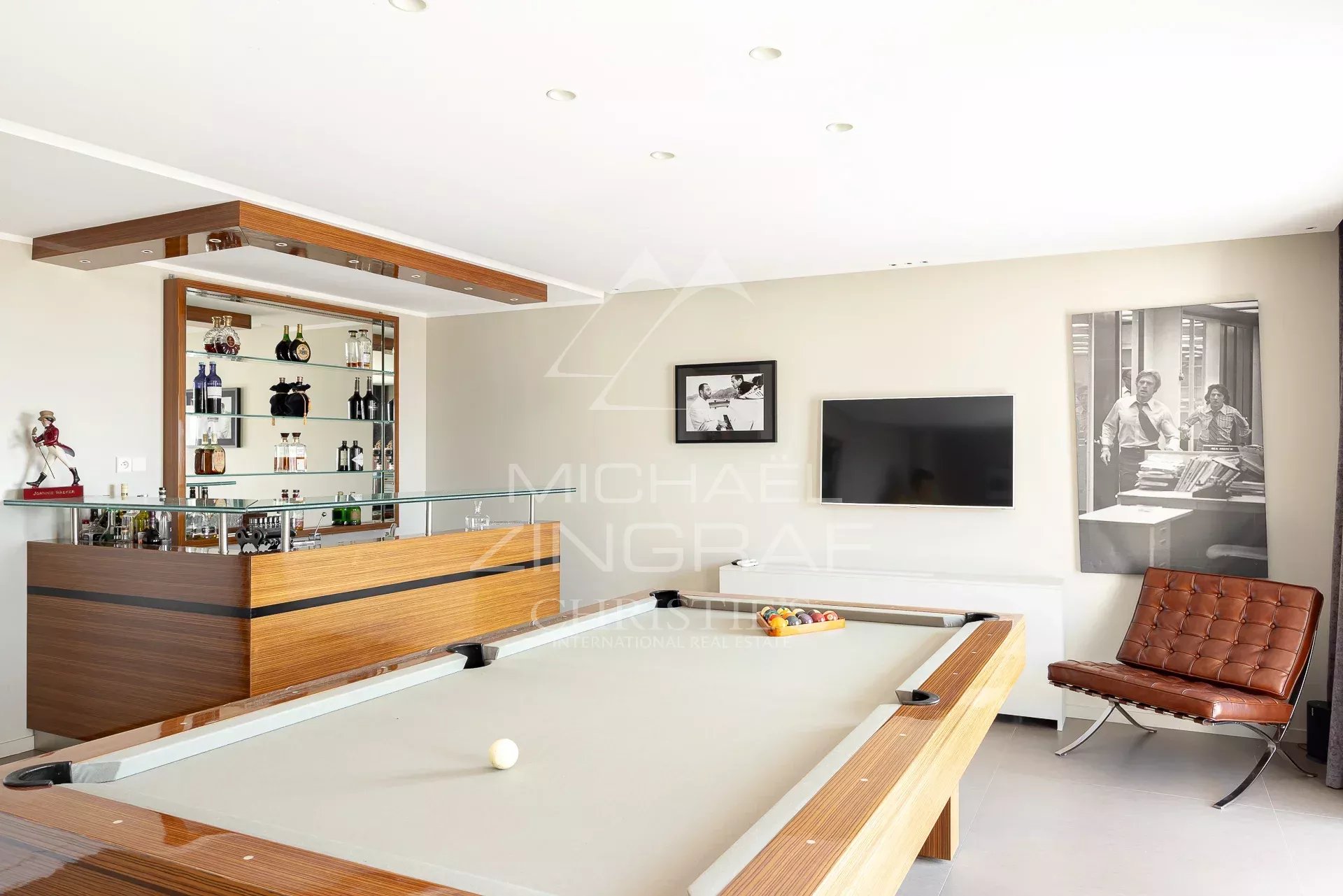 Contemporary home game room with a wooden bar, glass shelving of liquor bottles, and a pool table in the foreground. A brown leather lounge chair and wall-mounted TV are visible, with a black-and-white photo artwork nearby.