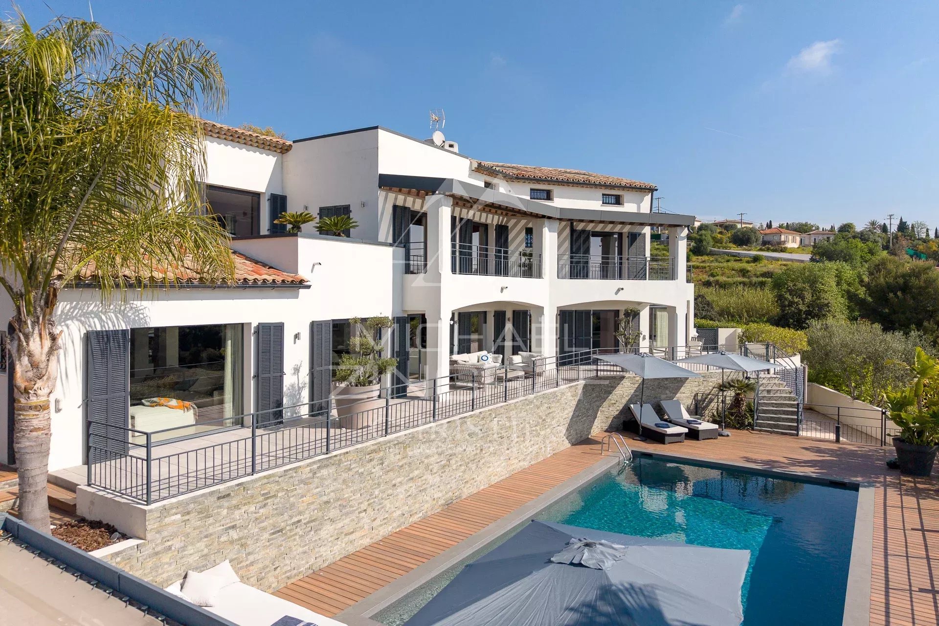 Luxurious white villa with two stories, multiple balconies, and a rectangular pool surrounded by a wooden deck and lounge chairs.