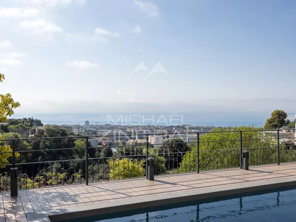 Poolside terrace with a black railing overlooking a coastal city and the sea under a clear blue sky, trees framing the scene.