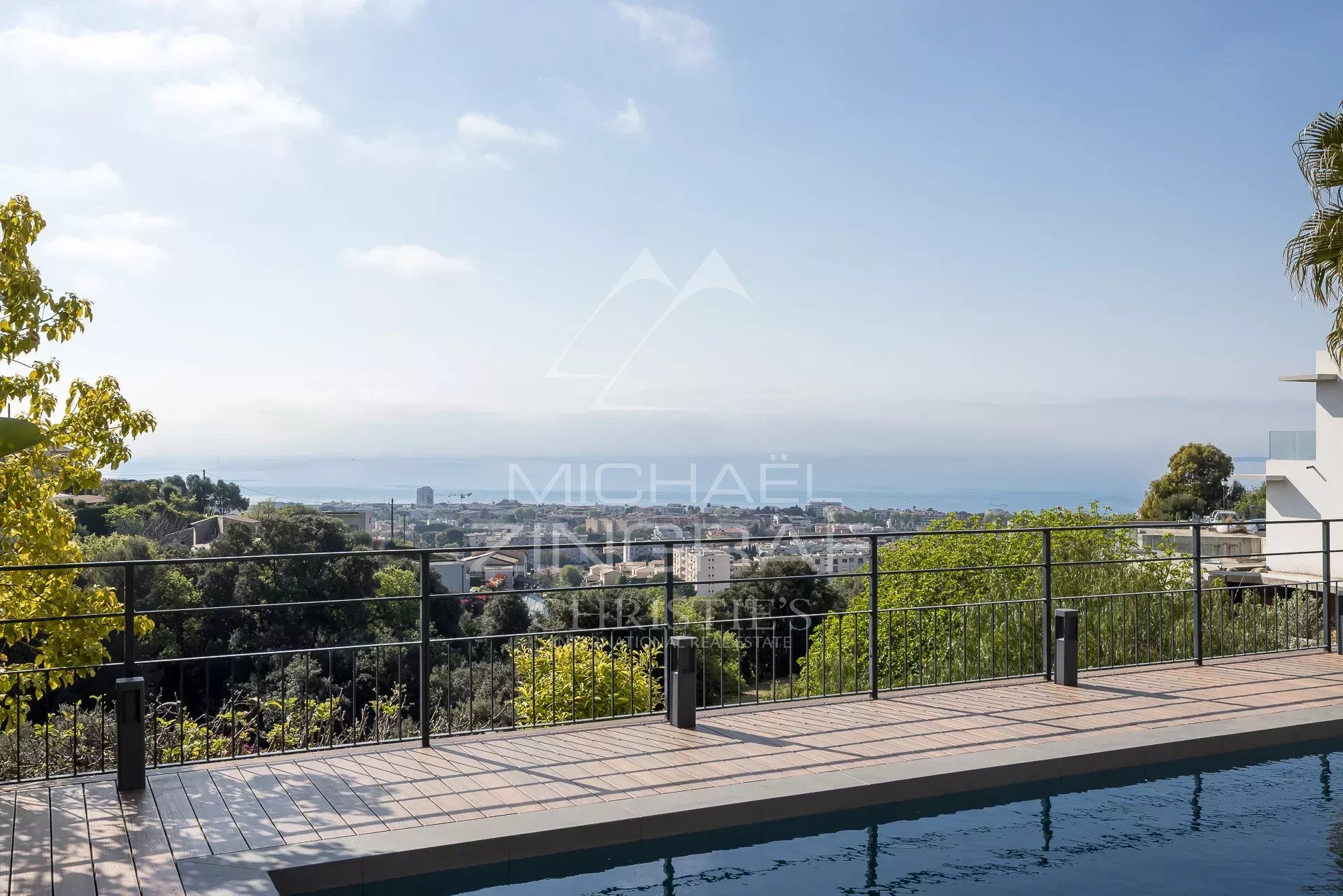 Poolside terrace with a black railing overlooking a coastal city and the sea under a clear blue sky, trees framing the scene.