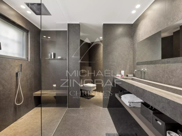Modern gray stone bathroom with a frameless glass shower, rainfall showerhead, and long countertop with dual sinks and open shelves beneath.