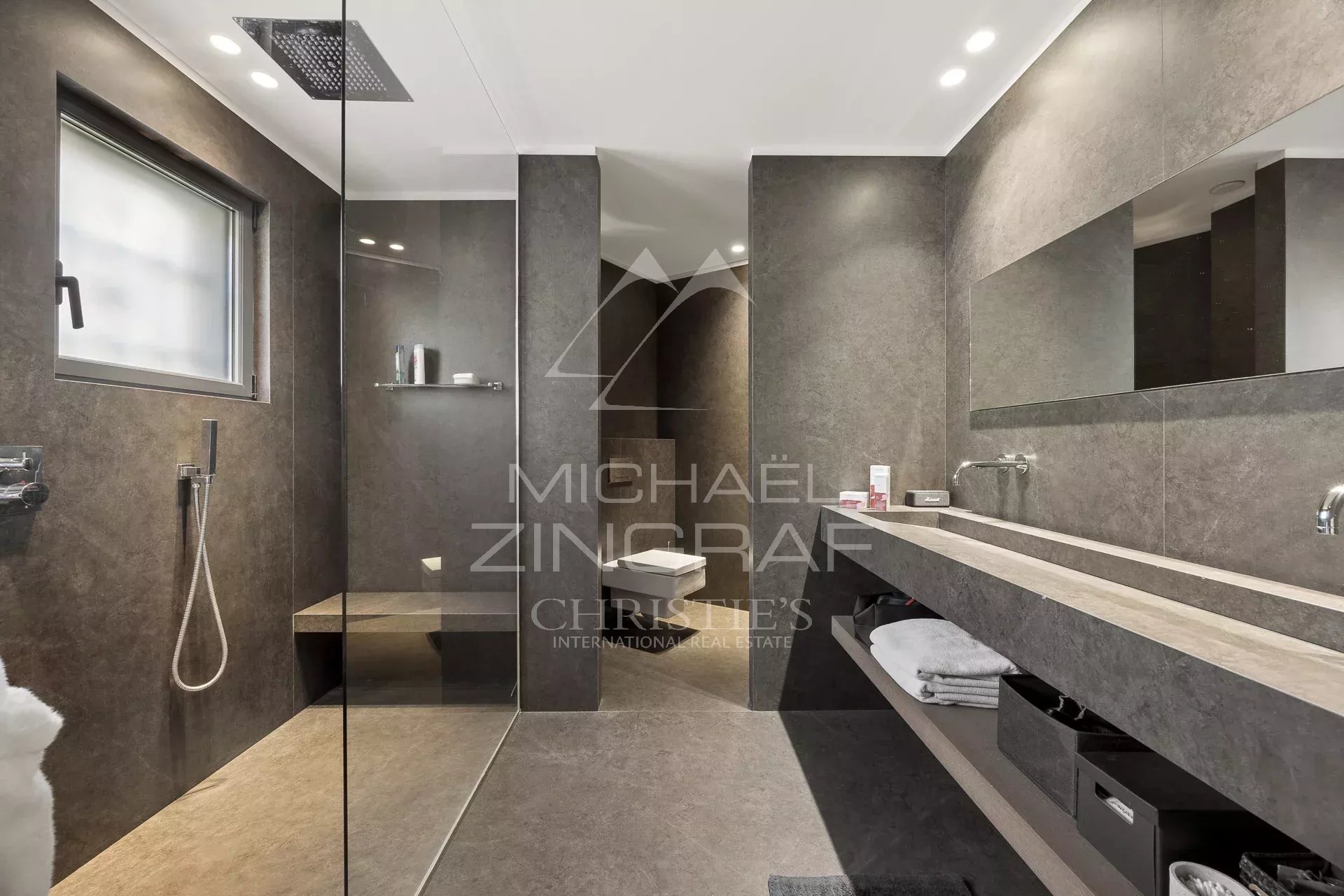 Modern gray stone bathroom with a frameless glass shower, rainfall showerhead, and long countertop with dual sinks and open shelves beneath.