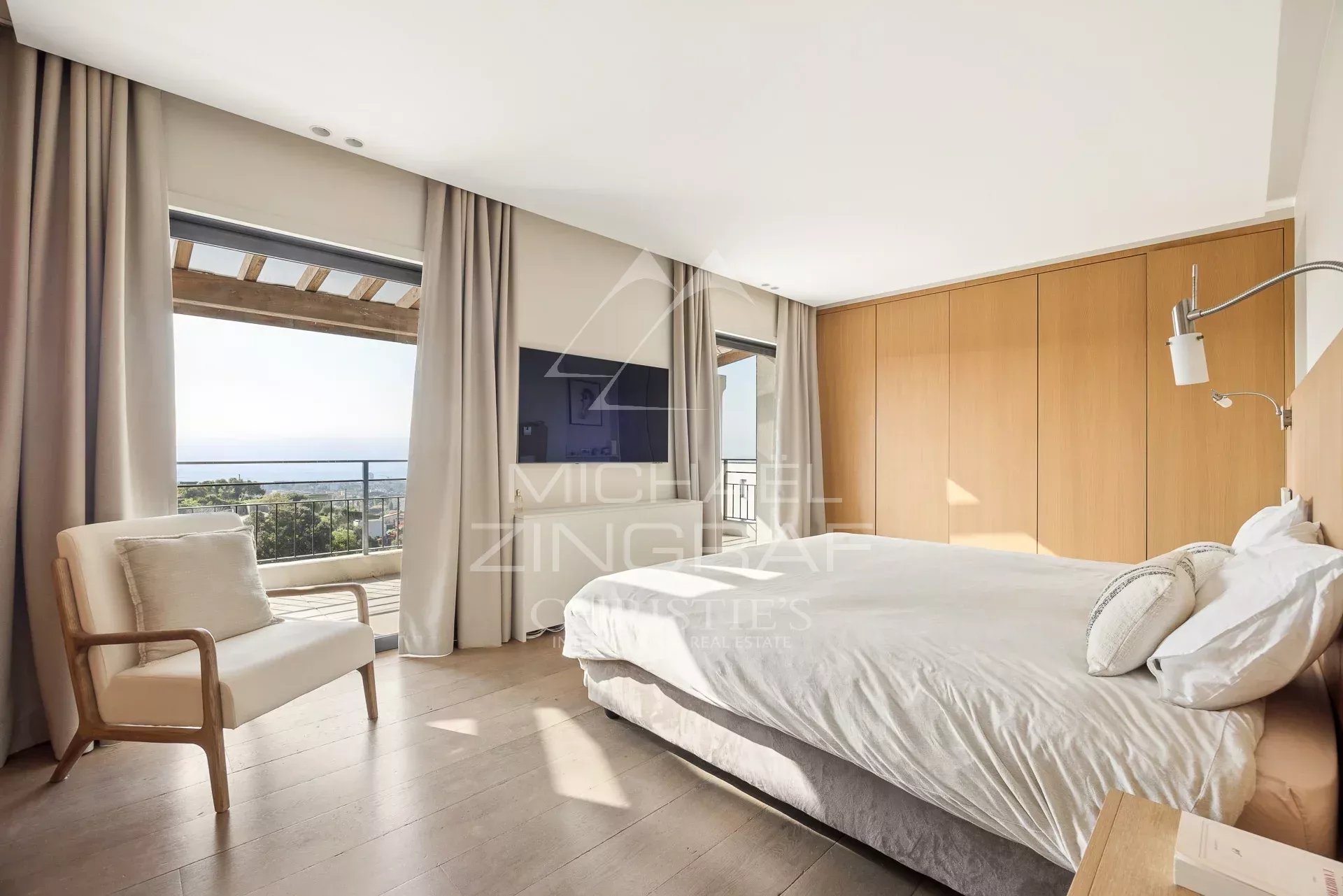 Bright hotel bedroom with a king bed, wooden wardrobe, and a balcony overlooking the sea from the window.