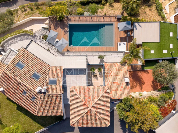 Top-down aerial view of a Mediterranean-style home with red-tiled roofs, a rectangular pool on a wooden deck, and surrounding greenery.