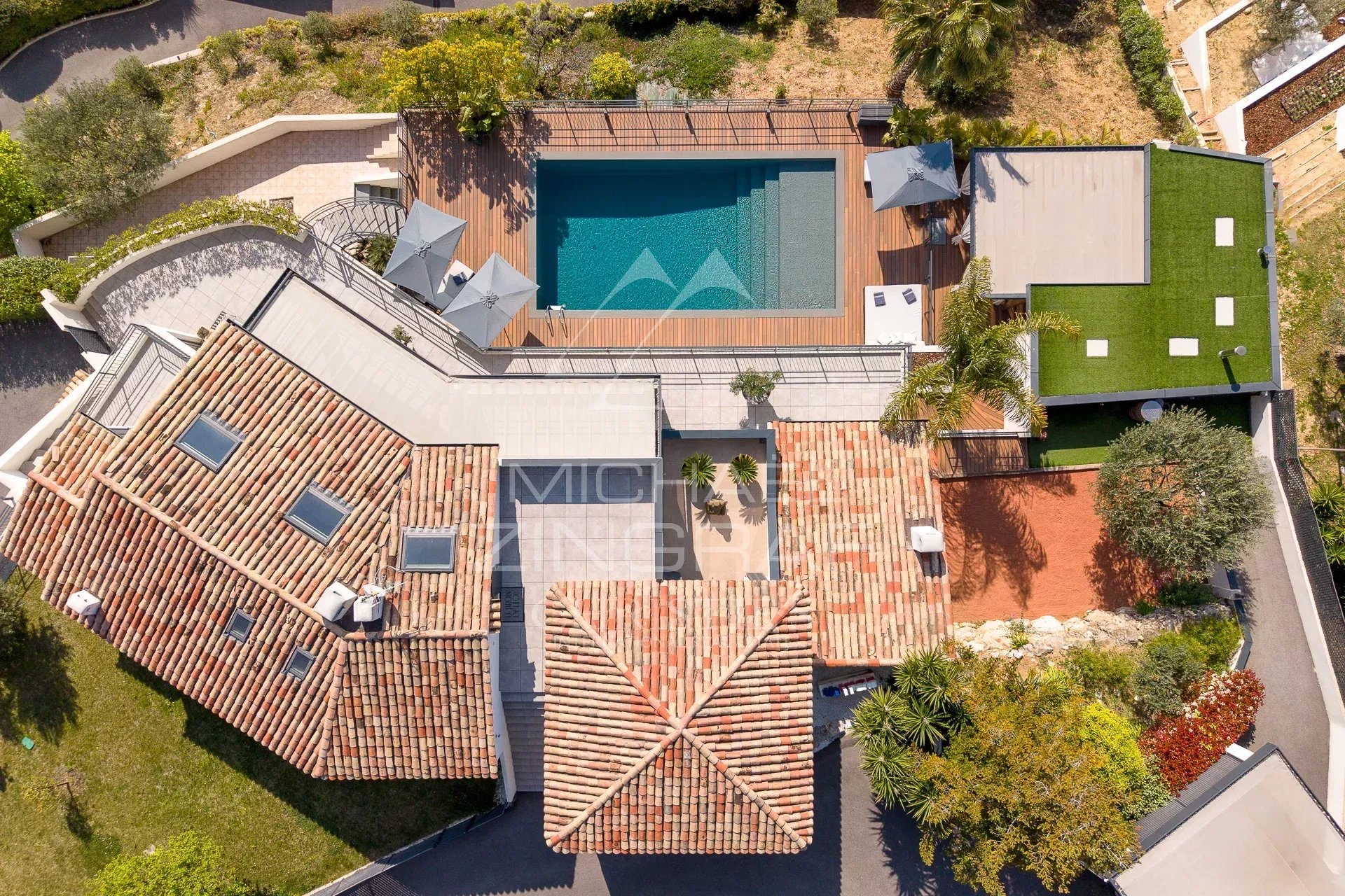 Top-down aerial view of a Mediterranean-style home with red-tiled roofs, a rectangular pool on a wooden deck, and surrounding greenery.