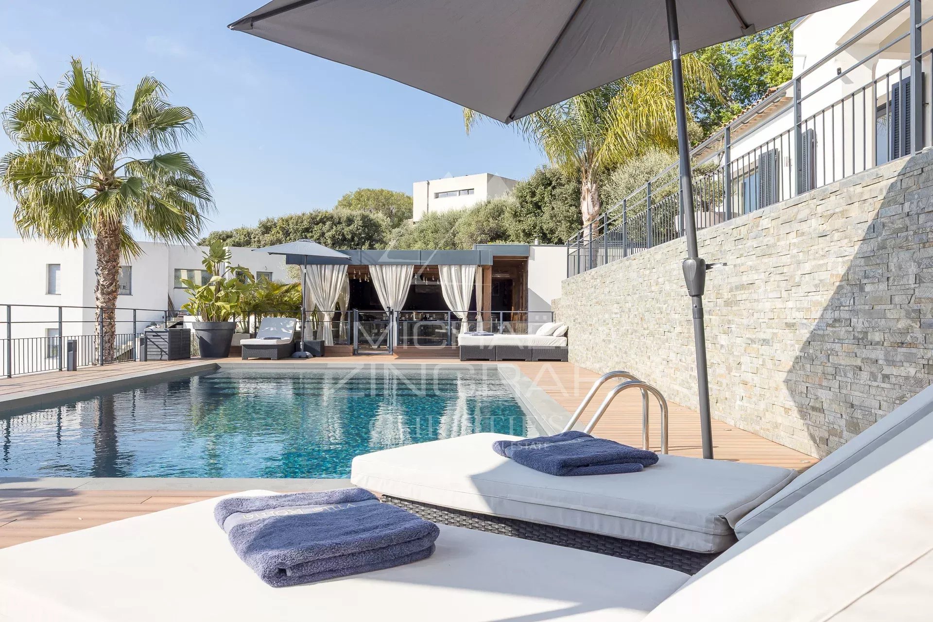 Poolside deck with white loungers, blue towels, and a large umbrella overlooking a rectangular pool and cabana.