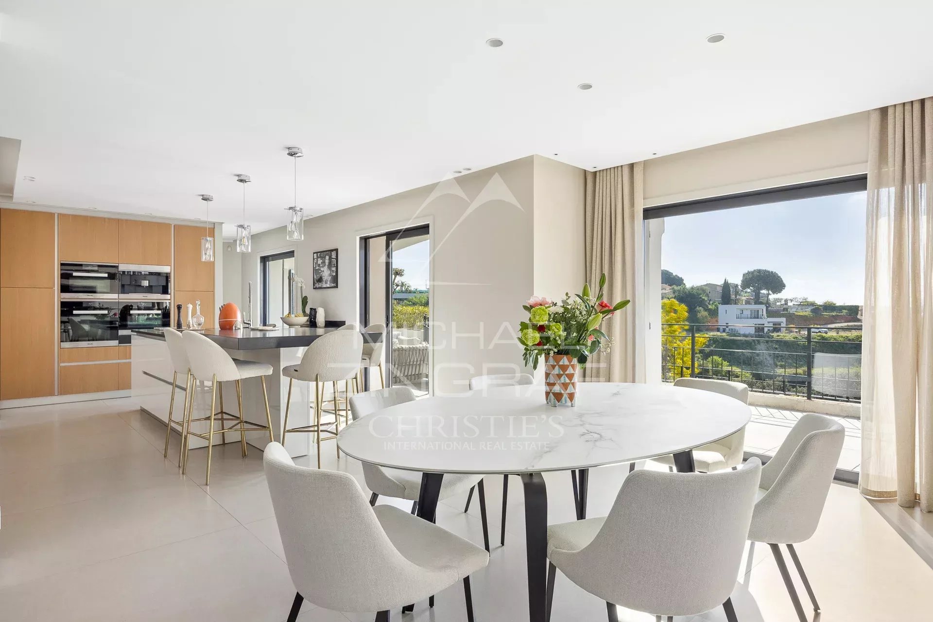 Bright open-plan kitchen and dining area with a round marble table, white chairs, and large windows leading to a balcony view.