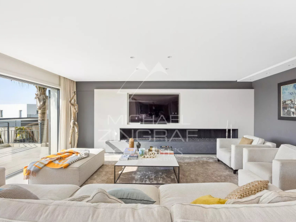 Bright modern living room with white sofas, a low glass coffee table, gray walls, and a balcony with an ocean view.